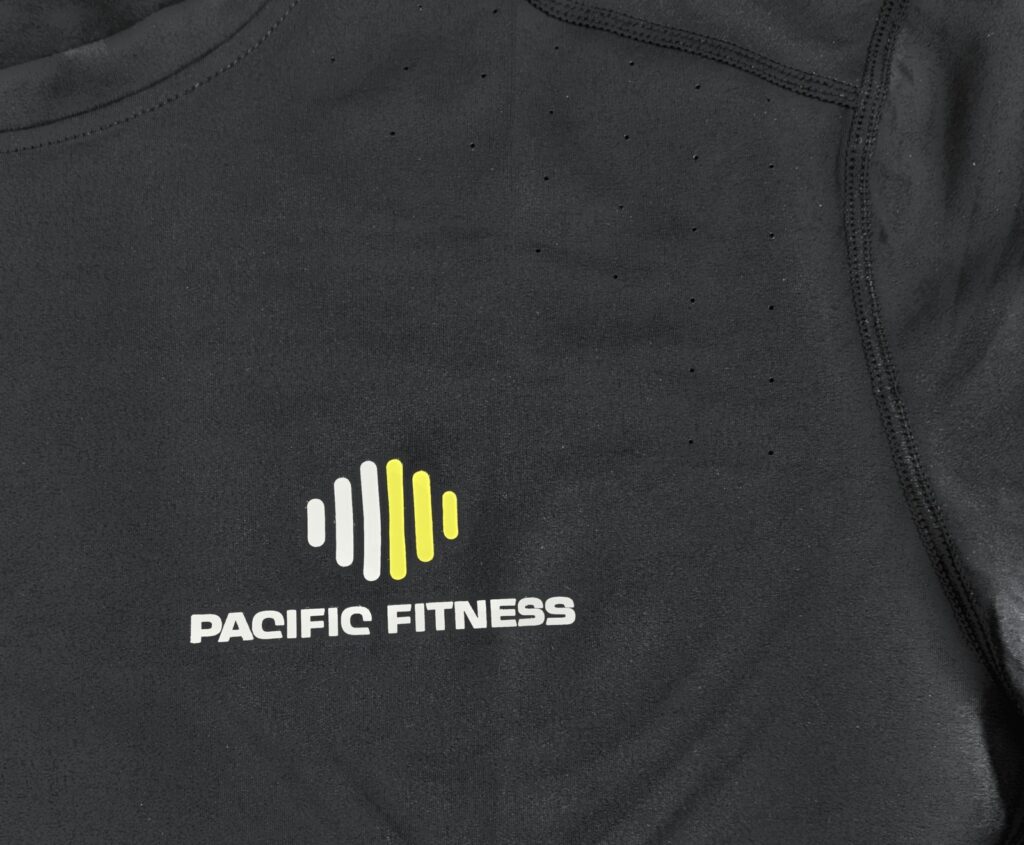 Gros plan du logo Pacific Fitness floqué en blanc et jaune sur un vêtement de sport noir à maille technique perforée, détouré sur fond clair.